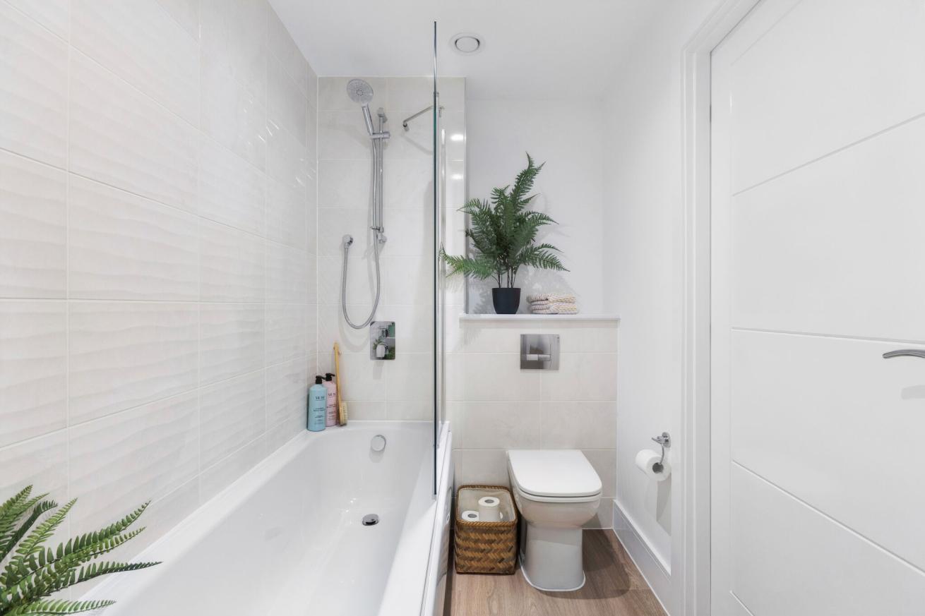 A modern, white bathroom with toilet and a shower over the bath, with decorative plants and a woven basket by the toilet