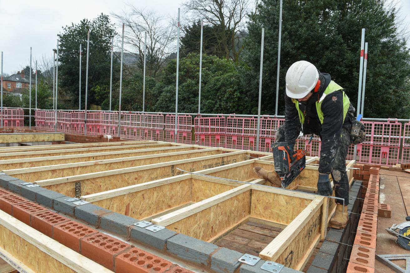 A construction worker wearing a hard hat and safety vest is using a nail gun to secure wooden beams on a building site. The framework consists of wooden planks and brickwork, with scaffolding and trees visible in the background. The area is surrounded by safety barriers.
