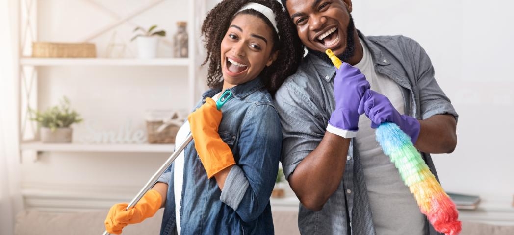 A young woman and man leaning on each other smiling and holding a duster and a brush