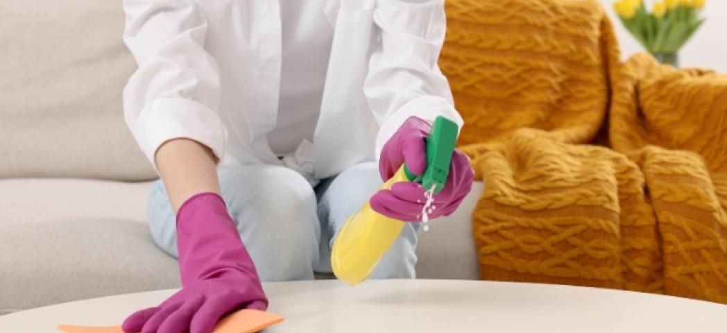 A person with yellow marigold gloves on holding cleaning spray and a sponge cleaning a wooden coffee table