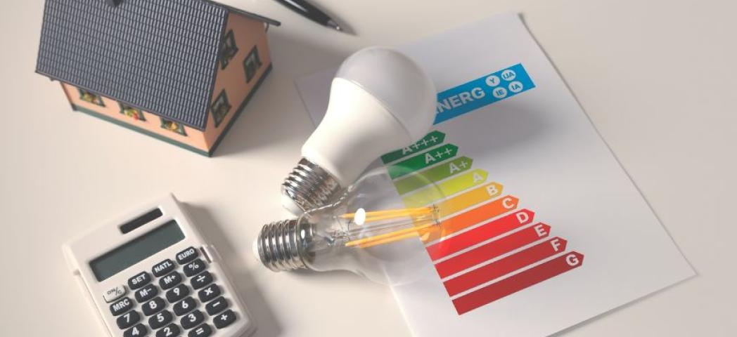 Centrally are two energy saving light bulbs resting on top of the energy efficiency paperwork, with a miniature house, standard calculator and pen scattered around them on a desk.