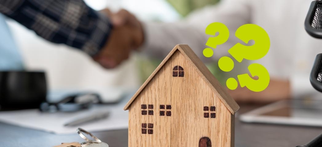 A wooden model of a house on a table next to a keychain with a silver key and a model house keyring on it. There are question marks around the model house and a handshake in the background