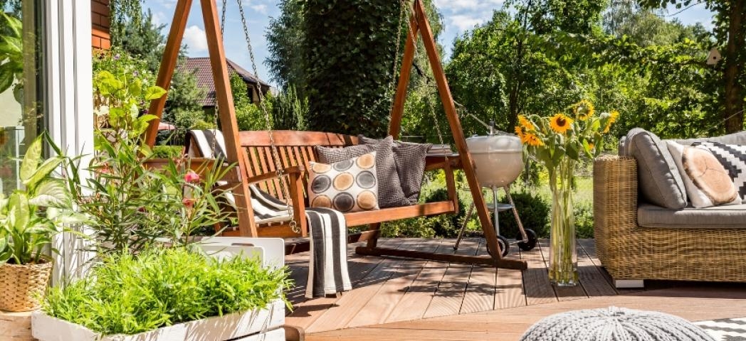 A wooden decking area with a swing seat, sunflowers, a barbecue and more plants 