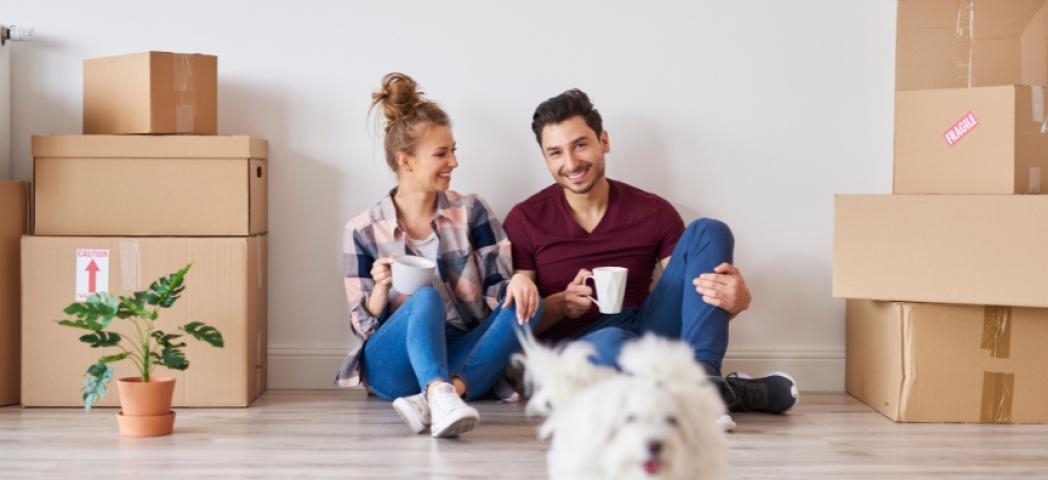 A couple sat on the floor of their new home with boxes beside them. They have a mug in their hand and look happy. Their small white dog is running towards the camera in front of them