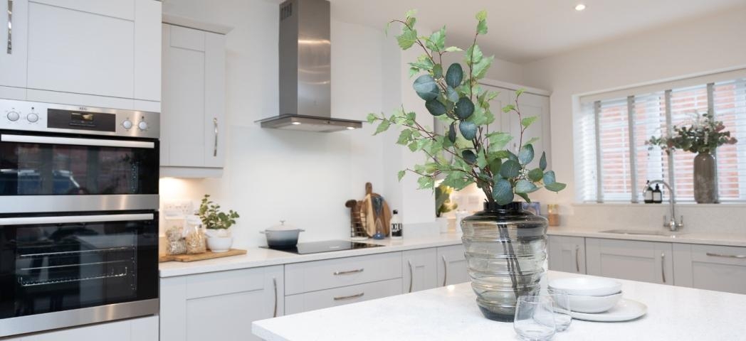 Interior of a clean and tidy white kitchen with cream cabinets. In the foreground is a kitchen island on which is a vase with foliage in and some crockery next to it
