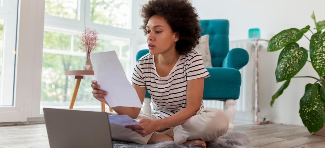 Stock image of a young woman sitting on a rug on a floor holding several pieces of paper and looking at a laptop