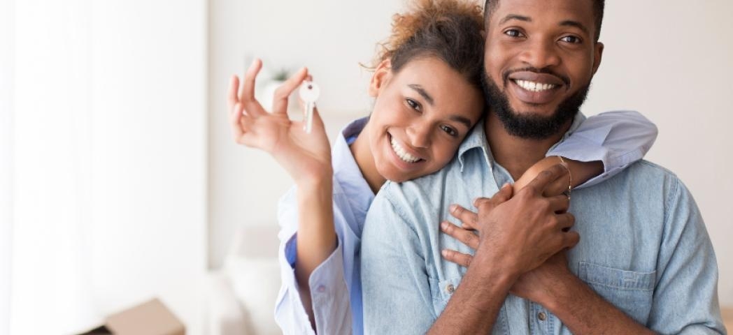 A male and female couple smiling at the camera, with the female holding up a key. She has her arm over the male in a sweet embrace