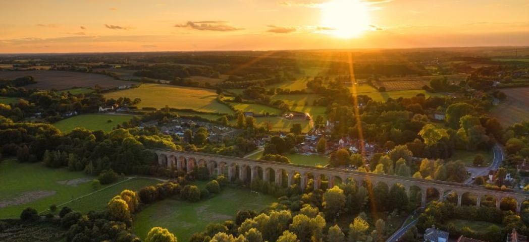 Aerial view of a countryside viaduct at sunset, with green fields, trees, and scattered houses.