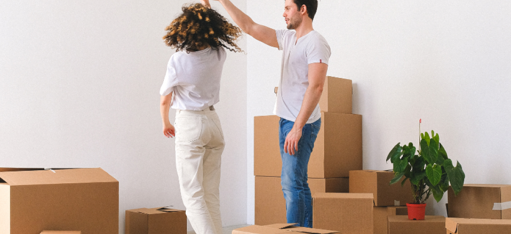 Two people in a room full of cardboard boxes, with one person playfully spinning the other by the hand. A green plant in a red pot is visible among the boxes.