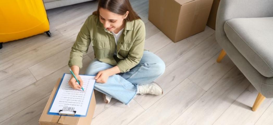 Woman sat on the floor and ticking off items from her moving day checklist while leaning on a closed box of possessions