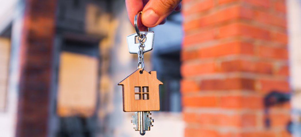 A house key with a home shaped wooden key ring attached are being held by two fingers. In the blurred background you can see a red bricked home.