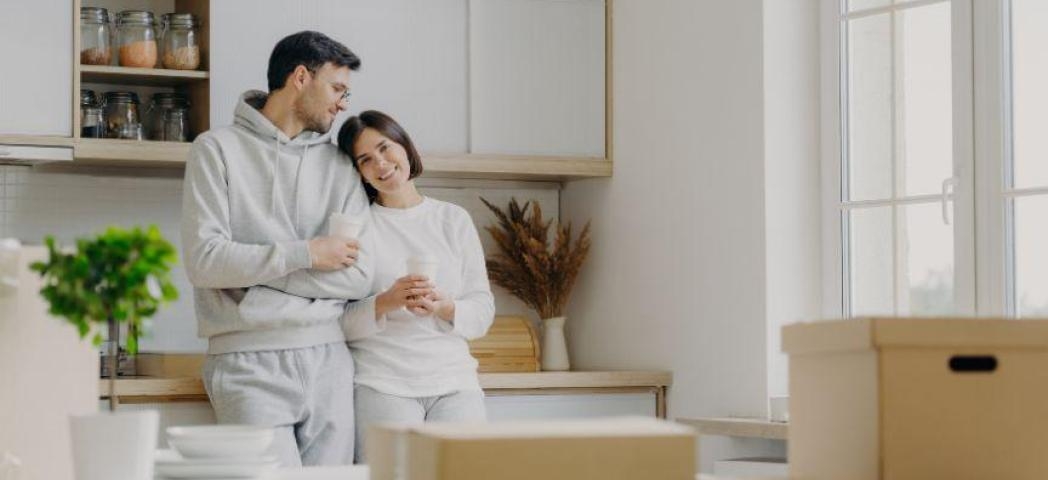A female stood leaning on a male against a kitchen worktop looking at moving boxes.