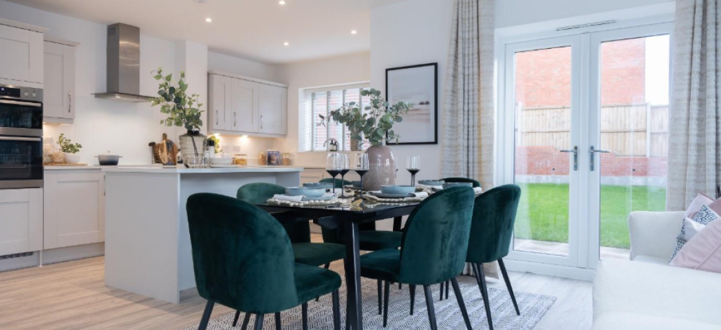 Open-plan kitchen/diner with white cabinets and quartz worktops. The dining space has a large table and 6 dark green velvet chairs, with a white sofa in the family area. Double doors open onto a rear garden.