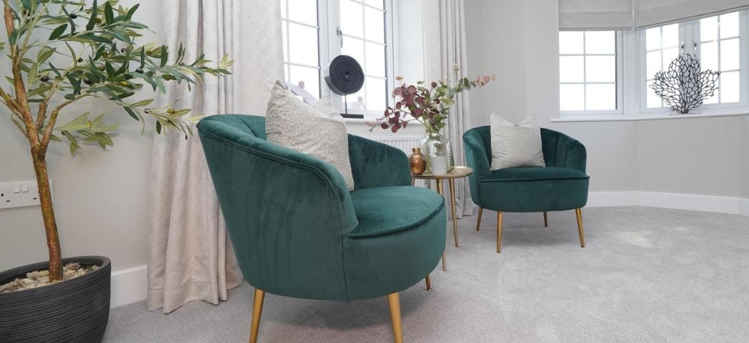 Two teal armchairs and a small side table in a bright, contemporary room with large windows, neutral décor, and a potted olive tree.