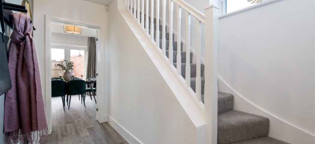 A hallway entrance to a home, with stairs and banister visible in the foreground and a dining room in the background