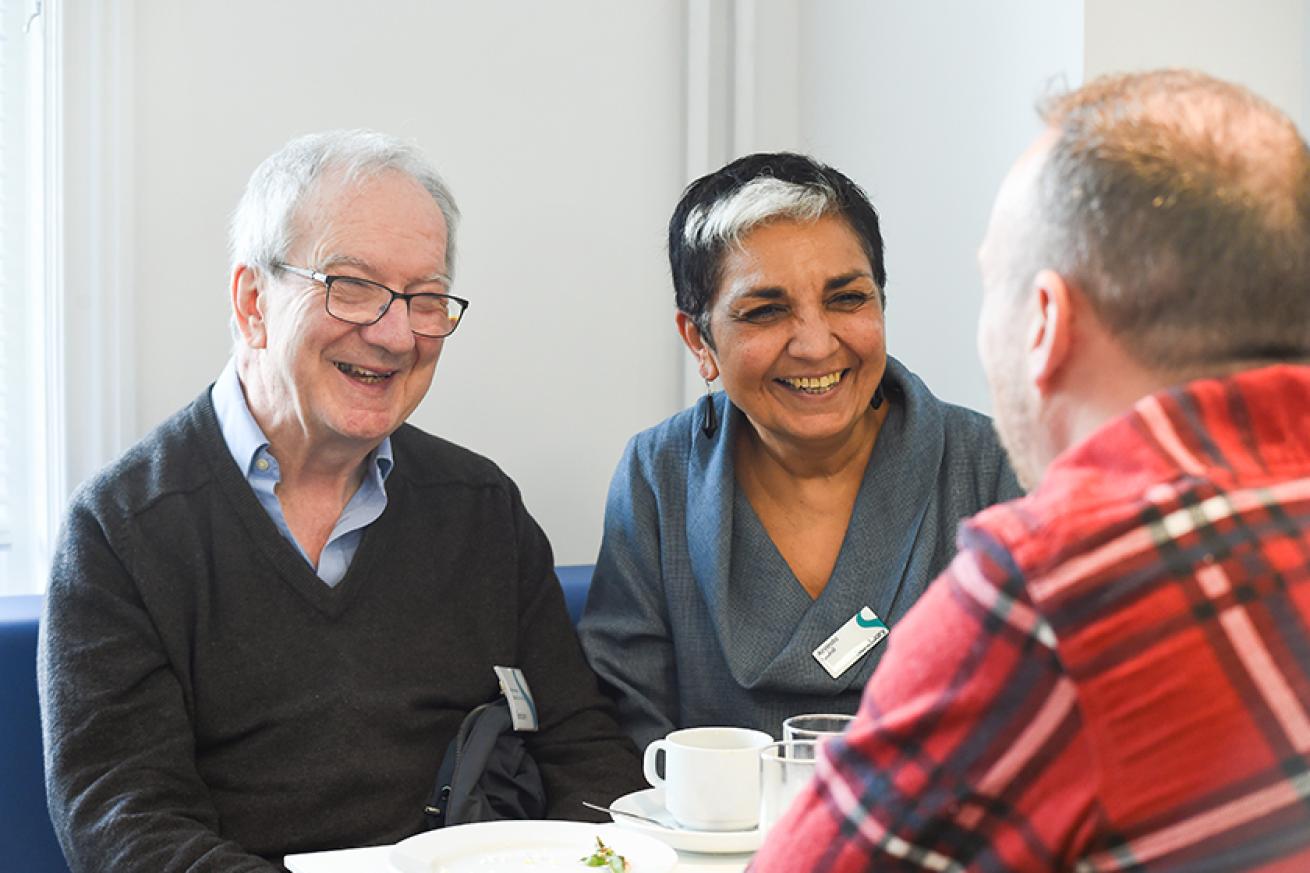 A woman with black and white hair wearing a grey top sitting on a blue bench next to a man wearing a blue shirt and grey jumper. Both people are wearing Sanctuary name badges and smiling at a man wearing a red checked shirt.