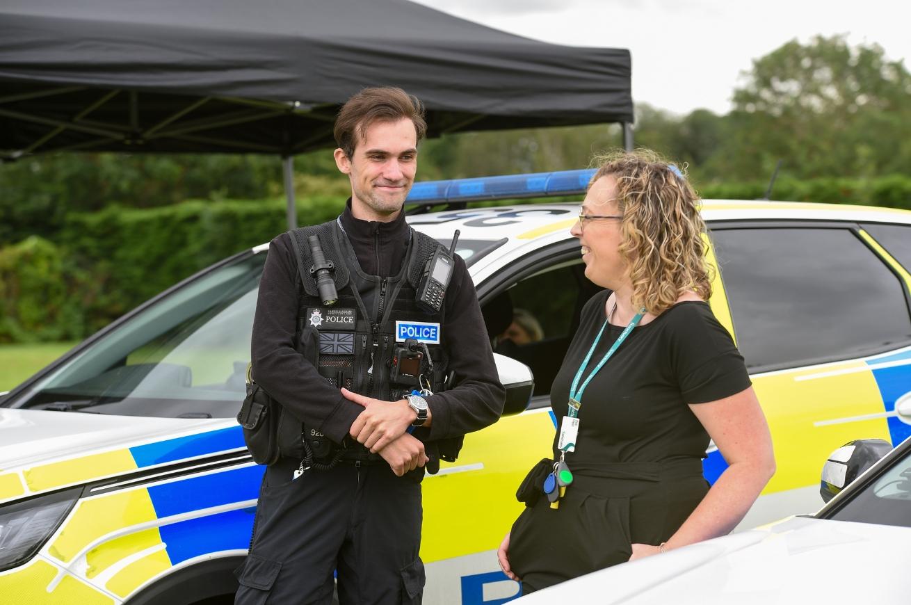 A police officer and Sanctuary employee stand in front of a police vehicle—one in uniform with gear, the other in casual clothes with an ID badge. They are having a conversation and smiling