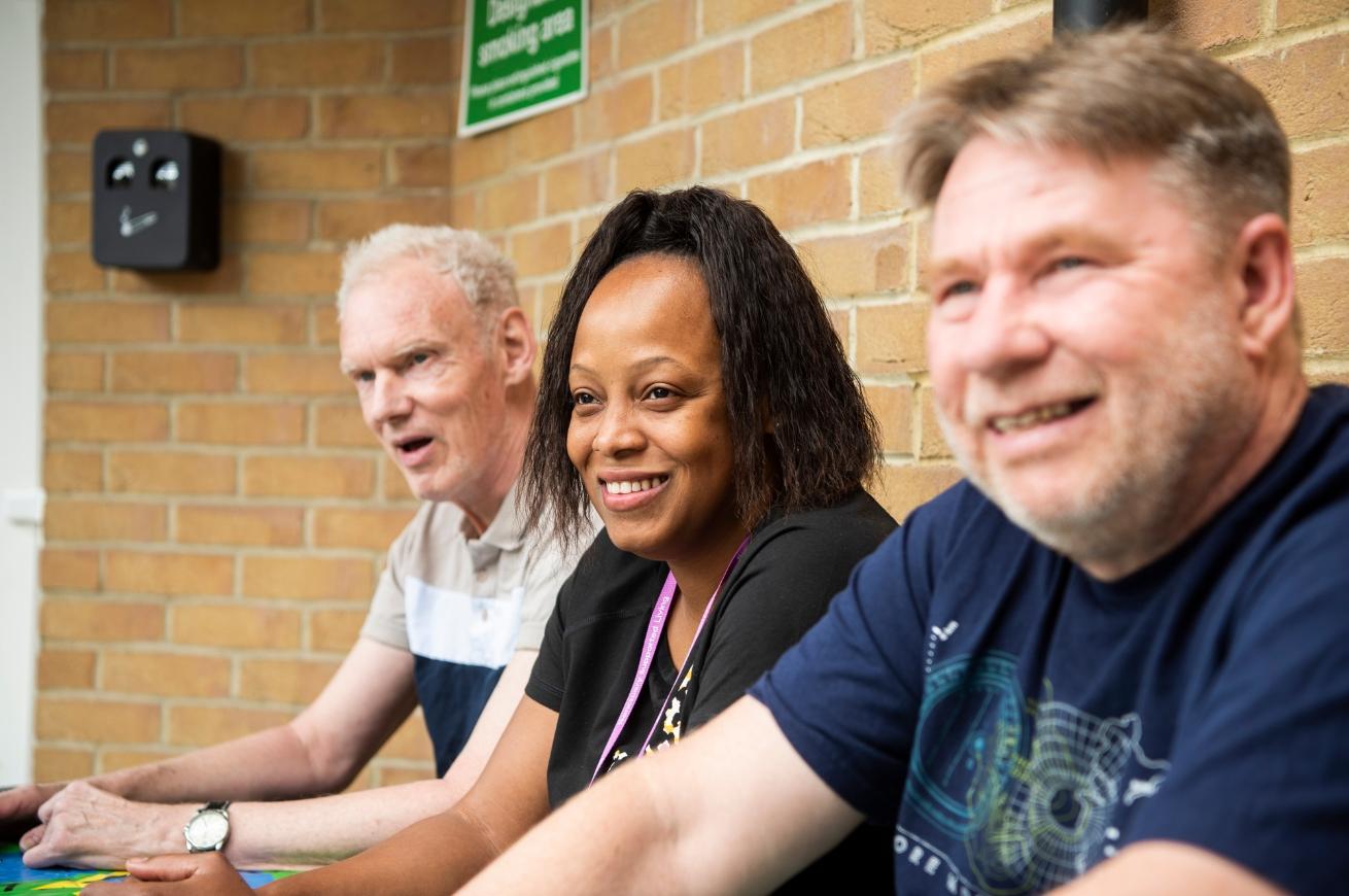Three people sit at a table against a brick wall, wearing casual shirts