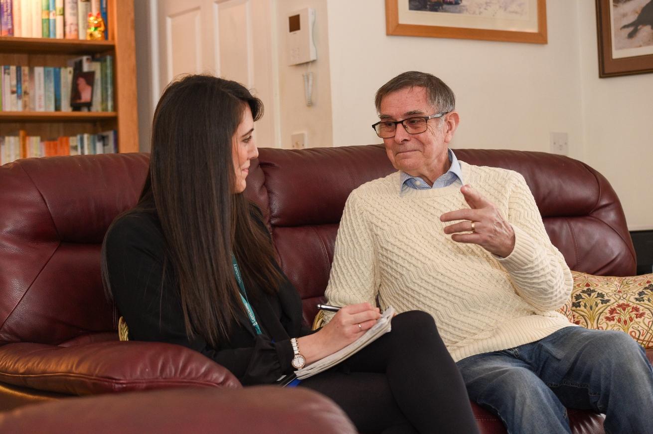 Two people sit on a maroon couch in a living room, one taking notes and the other gesturing. Bookshelves and framed pictures are in the background