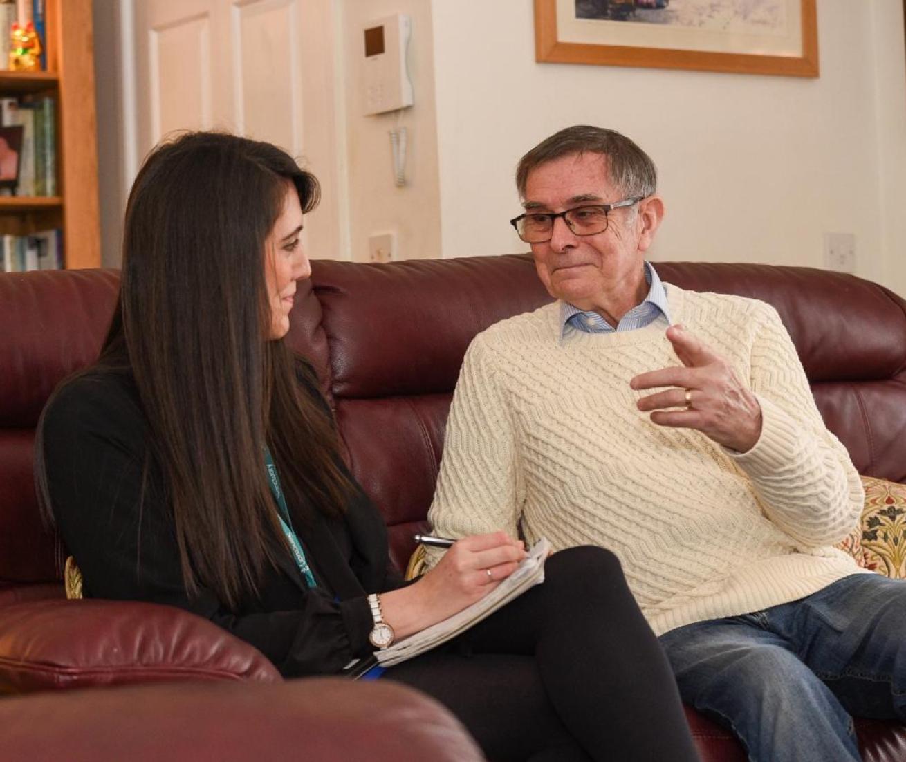 Two people sit on a maroon couch in a living room, one taking notes and the other gesturing. Bookshelves and framed pictures are in the background