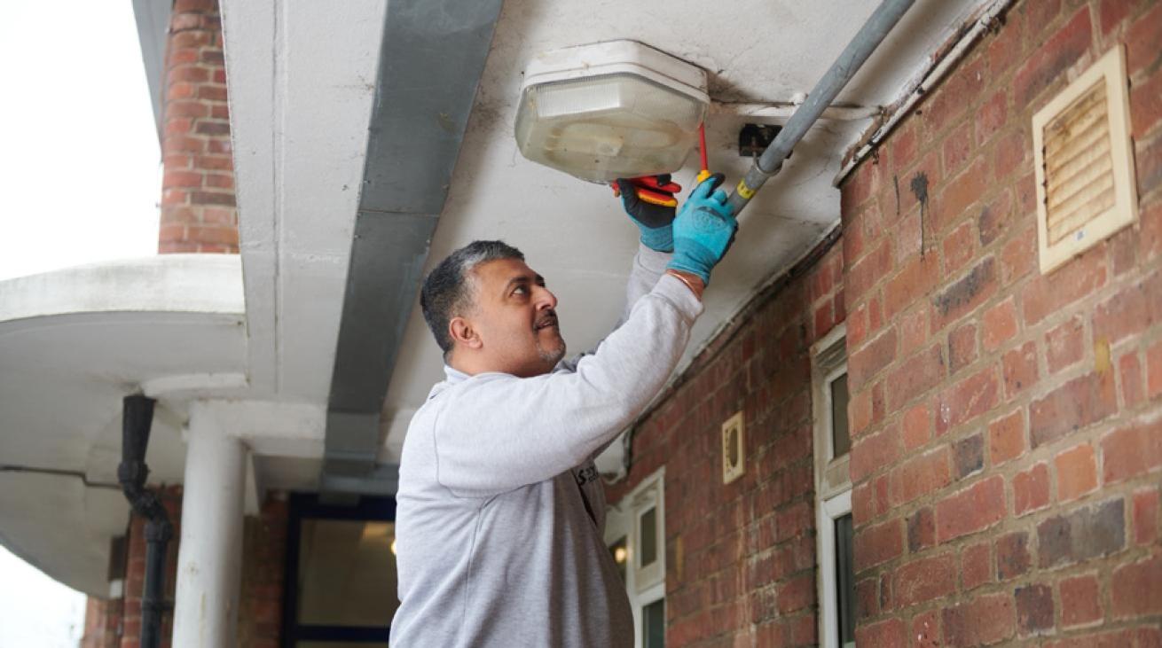 Person in gray sweatshirt and blue gloves repairing a ceiling light in a brick building