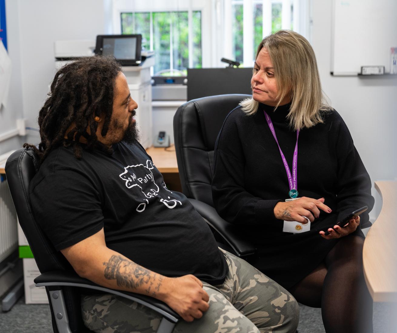 Two people sit in an office; one holds a tablet. The person on the left has dreadlocks and tattoos, the other wears a lanyard.