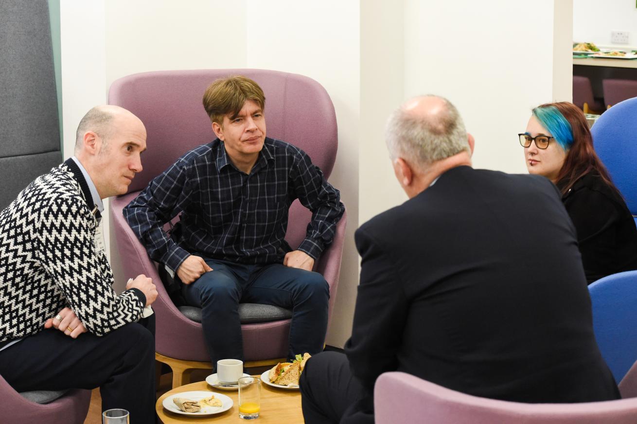 Four people sitting in a semi-circle on purple chairs, engaged in conversation. There are plates with food and drinks on the table in front of them