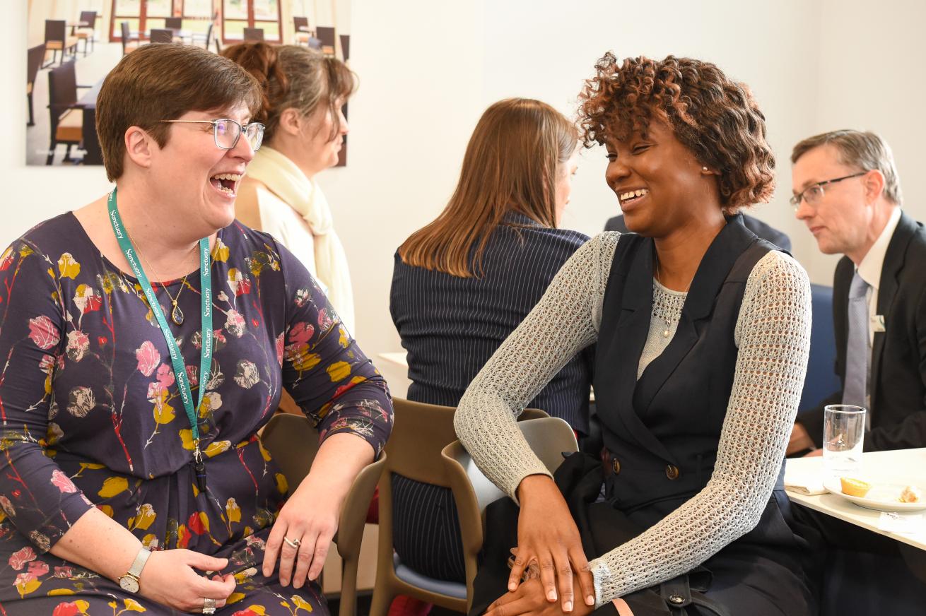 People engaged in conversation at a professional event, with two women in the foreground seated at a table, wearing name badges