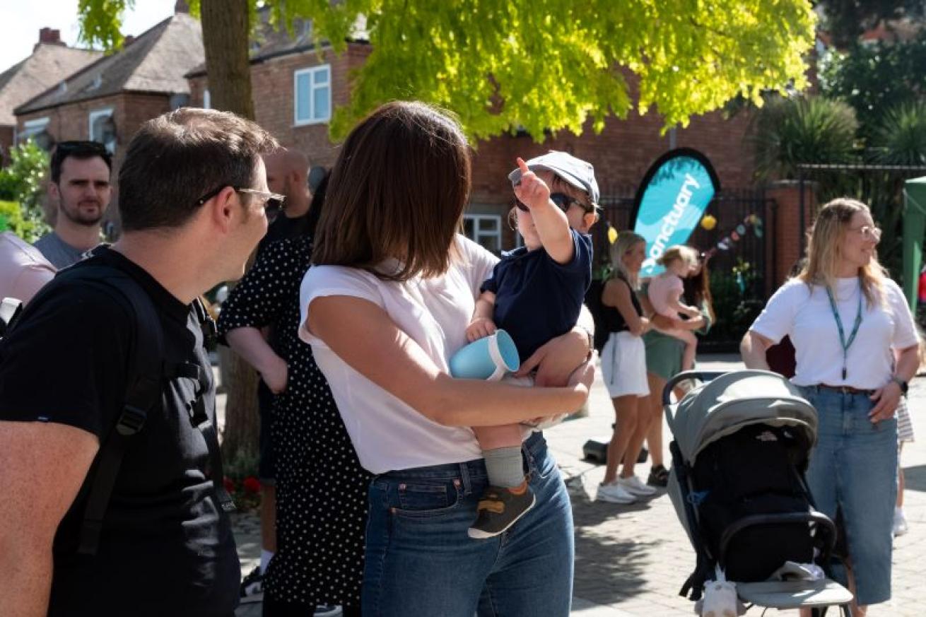 Sanctuary family fun day an outdoor gathering with families enjoying the sun; woman holding baby with blue sippy cup, stroller and others in the background.