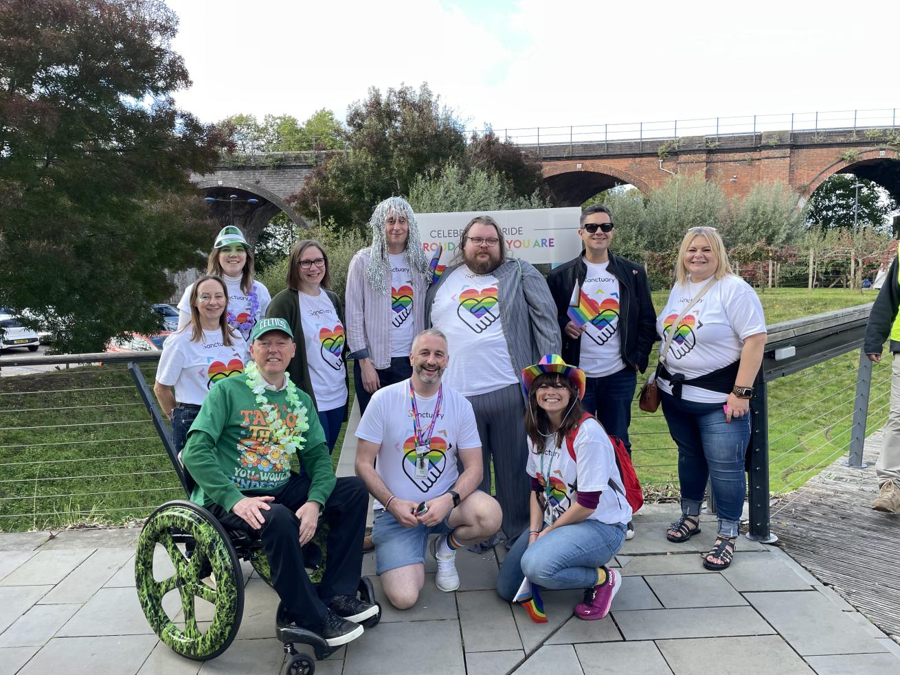 Group of people outdoors wearing pride-themed shirts, including one person in a wheelchair; greenery and a 'Celebrate Pride' sign in background.