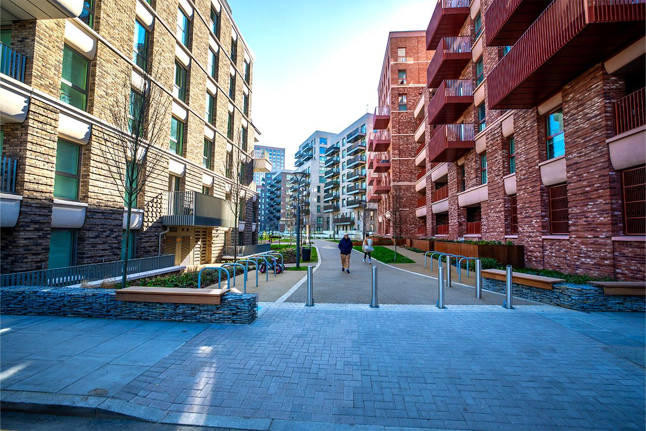 Two people walk along a tree-lined path between modern brick apartment buildings with balconies, bike racks, and benches in a sunny urban setting.
