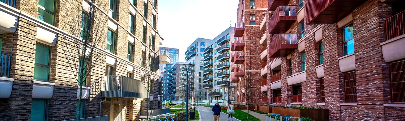Two people walk along a tree-lined path between modern brick apartment buildings with balconies, bike racks, and benches in a sunny urban setting.