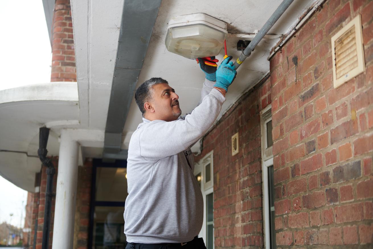A man wearing a gray sweatshirt and blue gloves is using tools to work on a light fixture mounted on a brick wall. He is smiling and appears focused on the task. The setting is an outdoor area with a covered walkway and a building in the background.