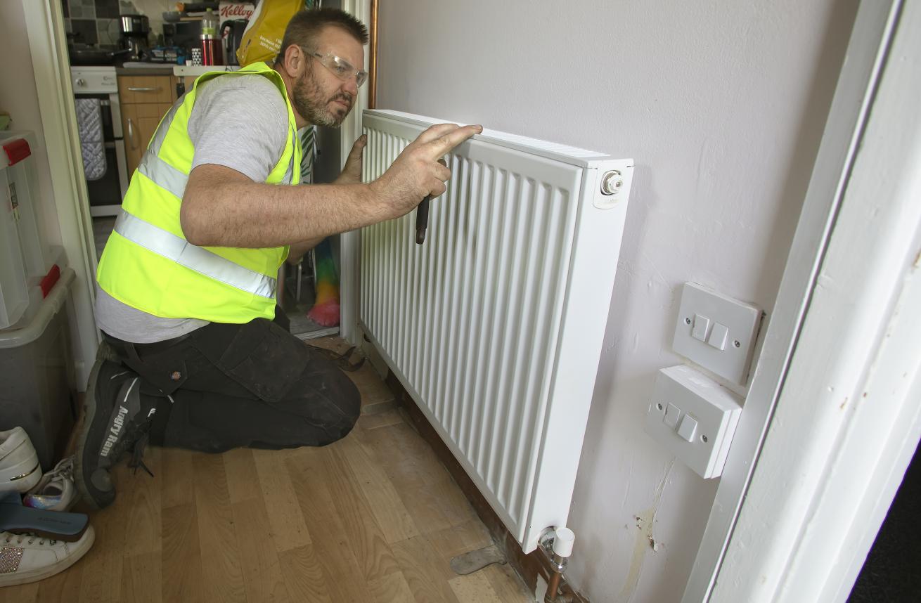 A maintenance worker in a high-visibility vest is kneeling beside a white radiator mounted on a wall. He is using a tool to adjust or repair the radiator. The background shows a kitchen area with various items visible, and there are light switches on the wall next to the radiator. The floor is wooden, and the overall setting appears to be a home or apartment.