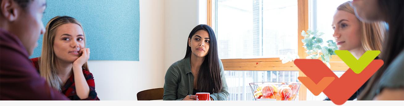 Five people sit around a table in a casual indoor meeting space with natural light. A bowl of fruit and assorted items rest on the table. 