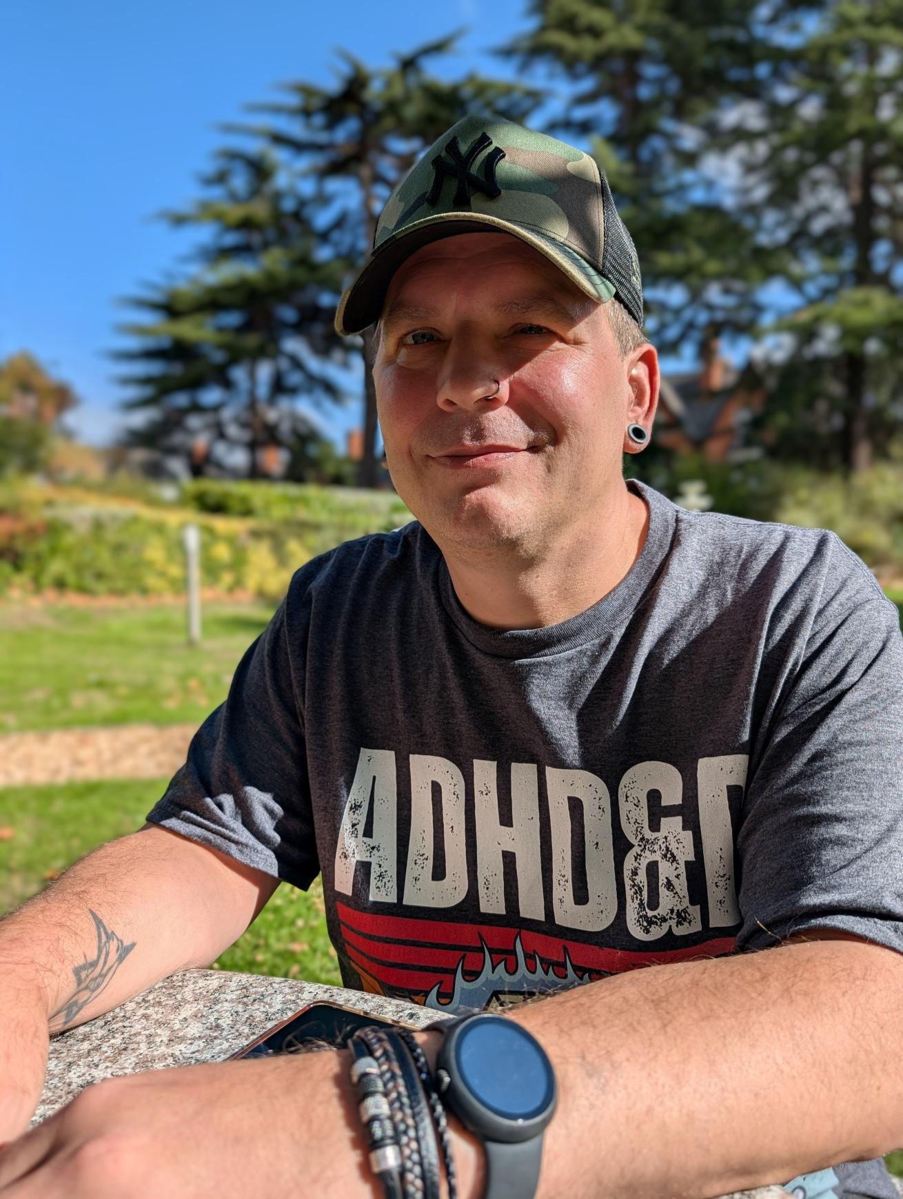 Sanctuary customer Alan Tyla looks towards the camera and smiles, He is sat outdoors in a grassy area and wearing a t-shirt and baseball cap.