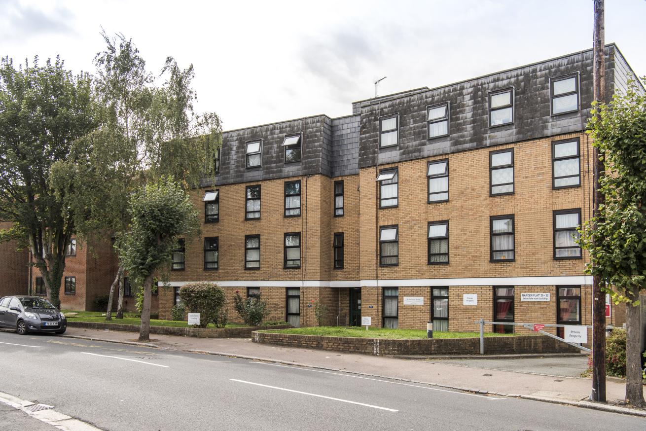 Modern multi-story brick building with flat roof and large windows, set along a tree-lined street with a parked car and sidewalk in view.