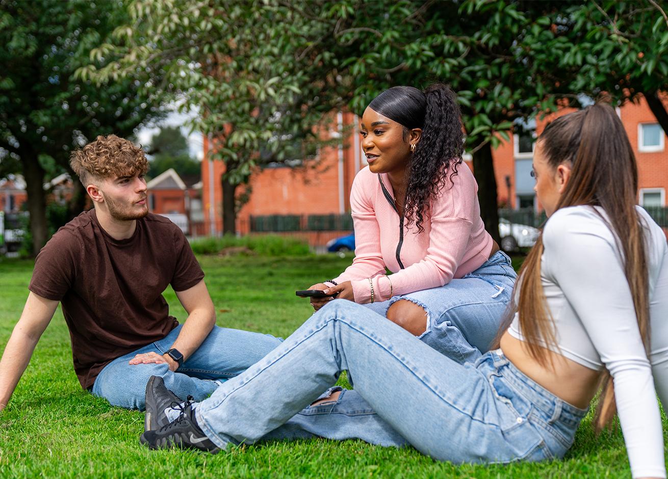 Three young adults are sitting on a grassy area in a park.