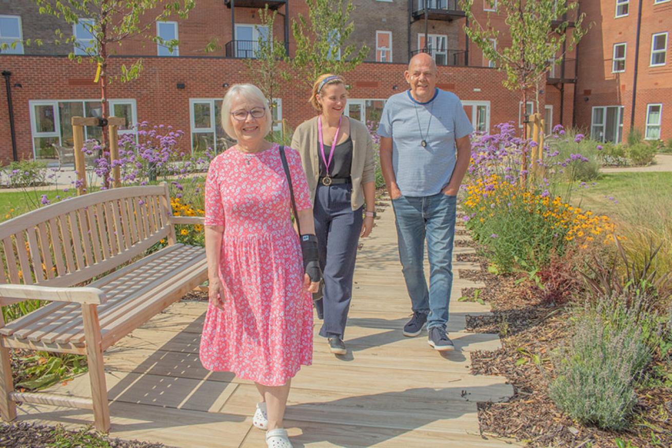 A sunny outdoor scene featuring three people walking along a wooden pathway in a garden. On the left, an older woman with glasses and short white hair wears a pink floral dress and white shoes, smiling at the camera. In the center, a middle-aged woman with brown hair, wearing a casual outfit and a lanyard, walks alongside the older woman. On the right, an older man with a shaved head and a casual gray shirt and jeans also walks, smiling. The garden is filled with colorful flowers and greenery, creating a vi