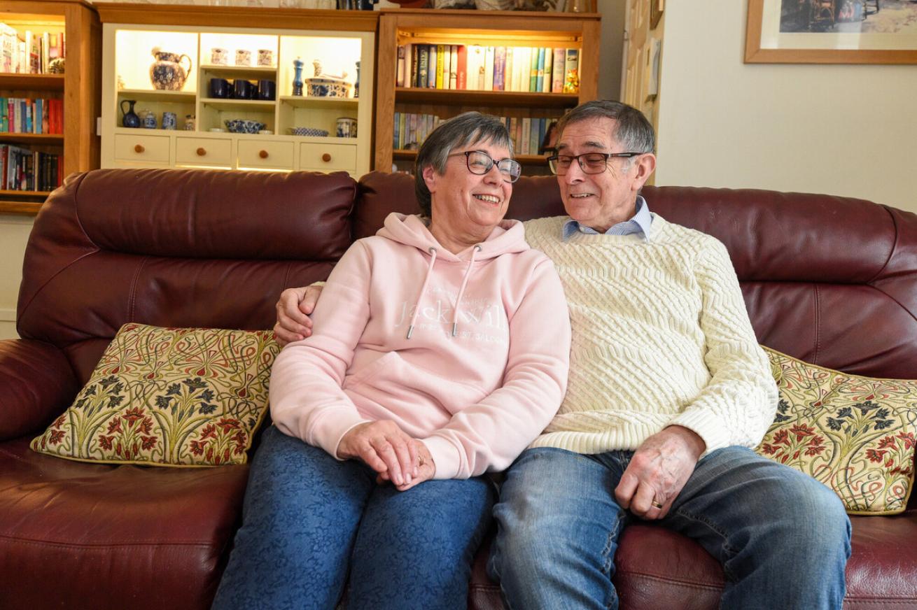 A middle-aged couple sits closely together on a comfortable sofa, smiling at each other. The woman wears a light pink hoodie and glasses, while the man is dressed in a cream-colored sweater and glasses. They are surrounded by a cozy living room with bookshelves and decorative items in the background. The atmosphere is warm and affectionate.