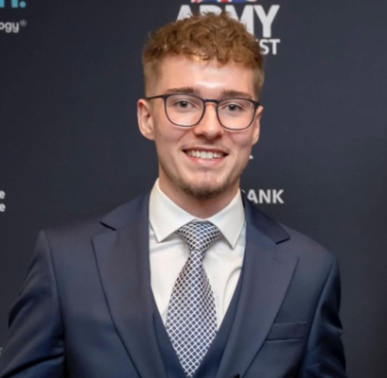 A man in a navy blue suit and patterned tie looks towards the camera and smiles. He has short brown hair and is wearing glasses.