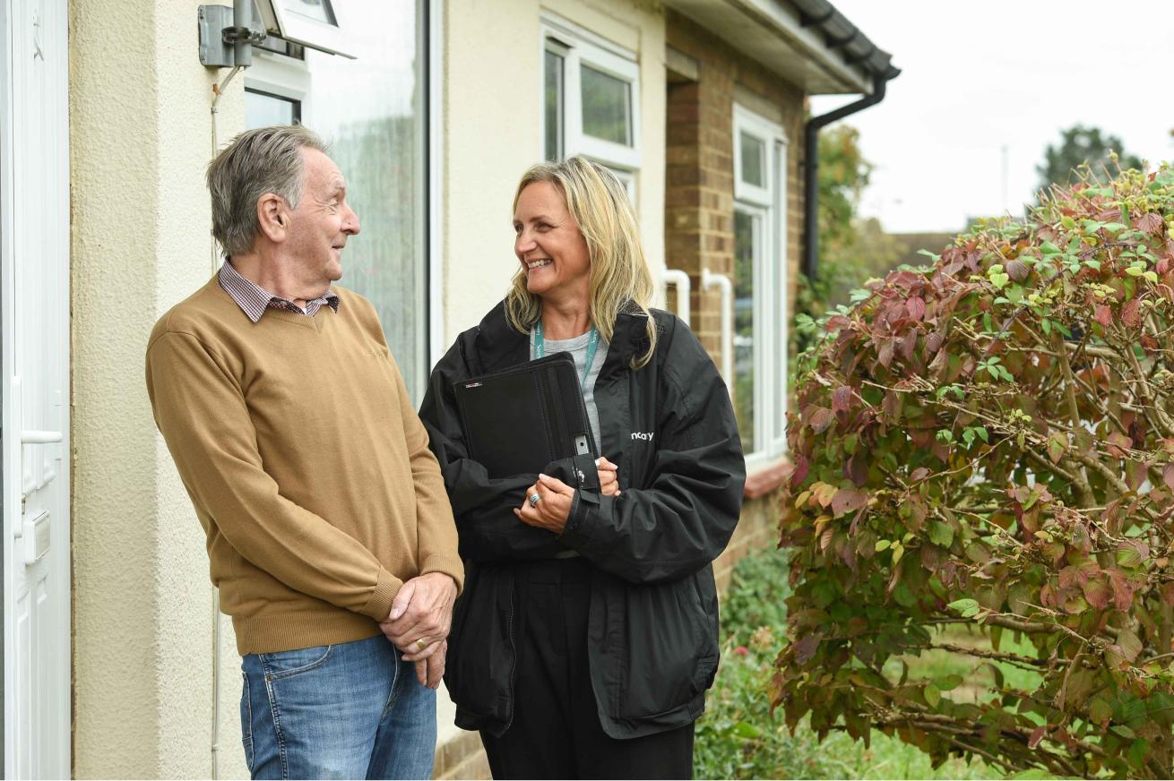 A man and a woman are standing outside a house, engaged in a friendly conversation. The man, wearing a brown sweater and blue jeans, is smiling at the woman, who is dressed in a black jacket and holding a clipboard. They are positioned near a bush with green and reddish leaves, and the setting appears to be a residential area with a cloudy sky in the background.