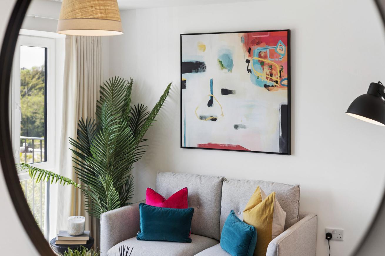 Modern living room corner featuring a light grey fabric sofa with colourful cushions in teal, mustard and pink, set against a white wall with a large framed abstract artwork. A tall green houseplant stands beside floor-length cream curtains and a glass door letting in natural light. A black wall-mounted lamp and a woven pendant light add warm accents, while a small side table with books and a candle completes the contemporary, styled space.