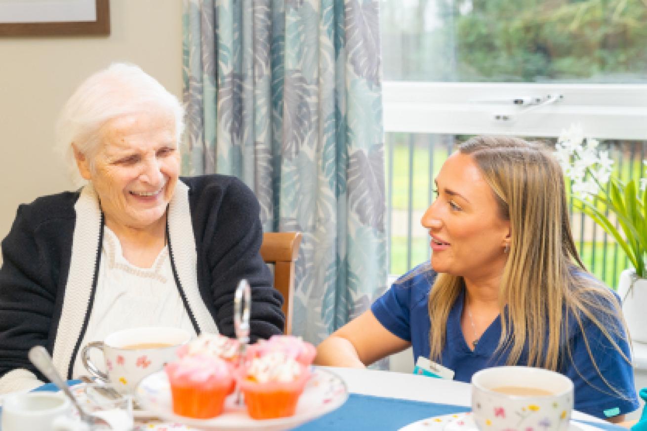 An elderly woman with white hair and a warm smile sits at a table, laughing with a younger woman in a blue uniform.