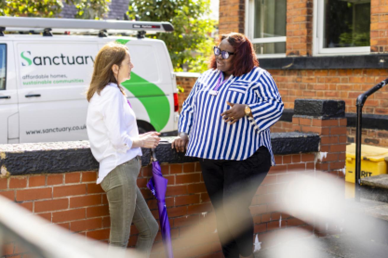 Two women are engaged in conversation outside a building. One woman, wearing a striped blue and white shirt, stands with her arms crossed, smiling. The other woman, dressed in a light-colored top and jeans, holds an umbrella.