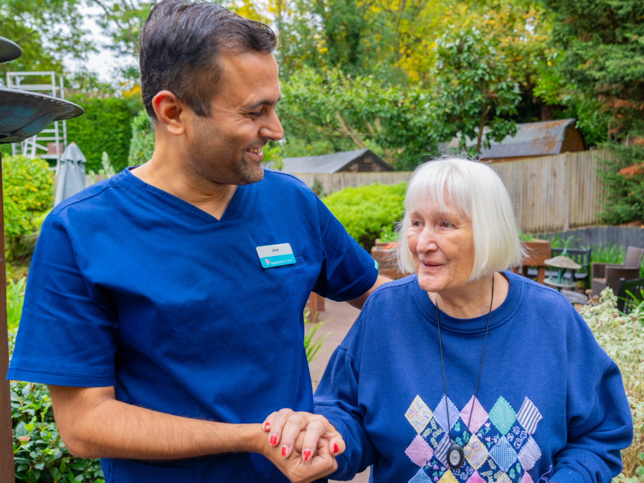 A healthcare worker in a blue uniform smiles and holds hands with an elderly woman in a blue sweater adorned with a diamond pattern