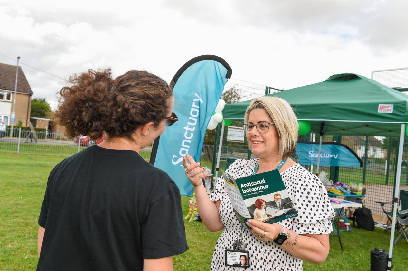 A woman with short blonde hair and glasses is speaking to a person with curly hair, wearing a black shirt and sunglasses. The woman is holding brochures titled "Antisocial behaviour" and is gesturing as she talks. In the background, there is a green tent with the word "Sanctuary" displayed on a banner, and various items are set up on a table. The setting appears to be an outdoor community event, with a grassy area and a fence in the background.