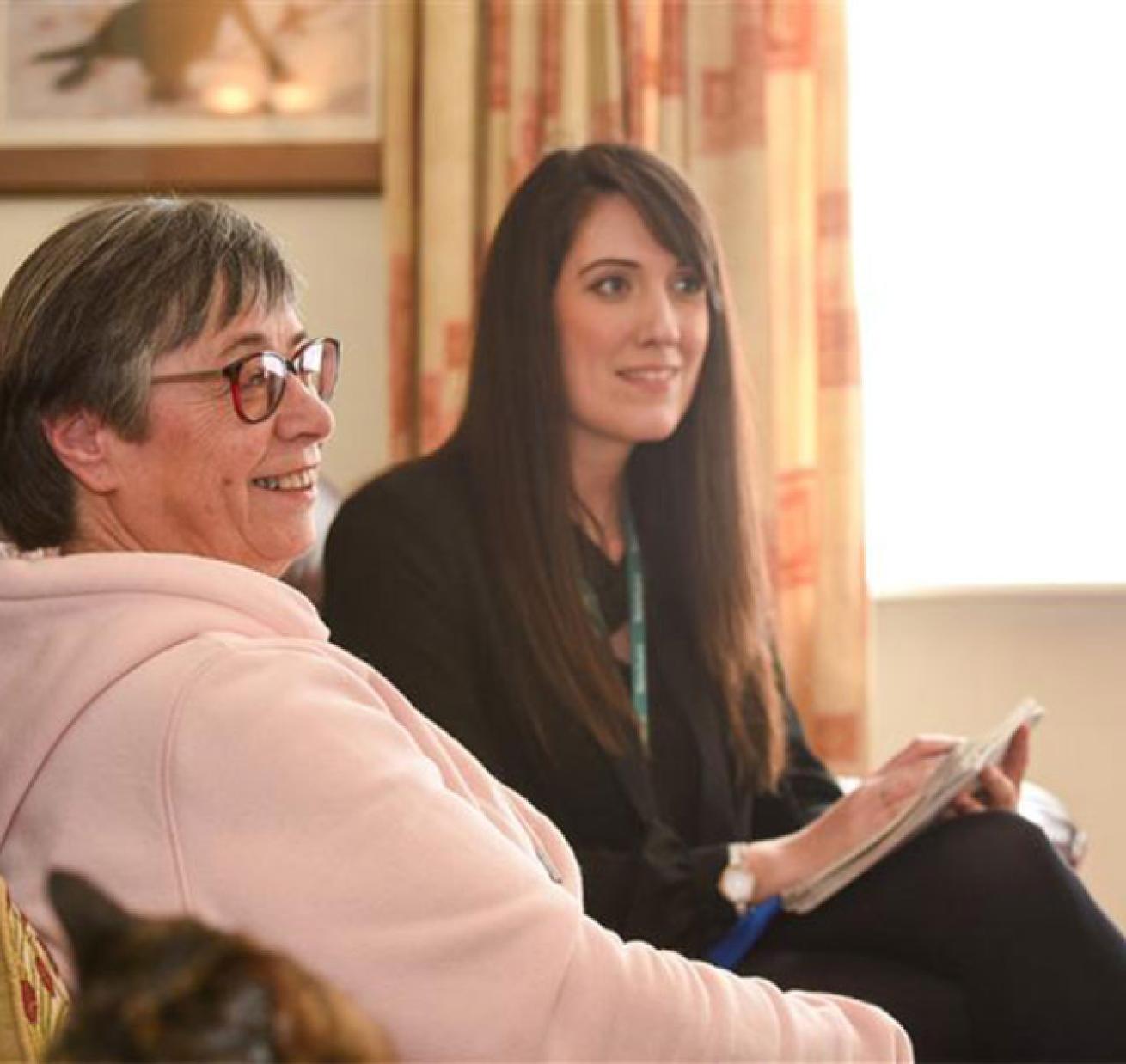 Two people sitting indoors, talking, with one person holding a notebook.