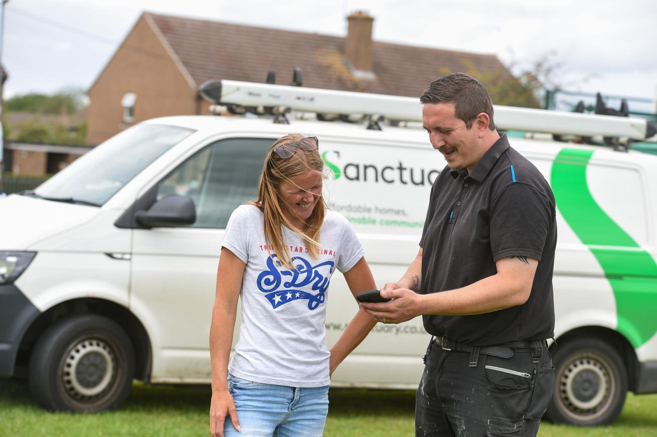 Two people stand outside on a windy day in front of a Sanctuary maintenance van. Sanctuary repairs staff member is showing a resident something on his phone, they are both smiling.