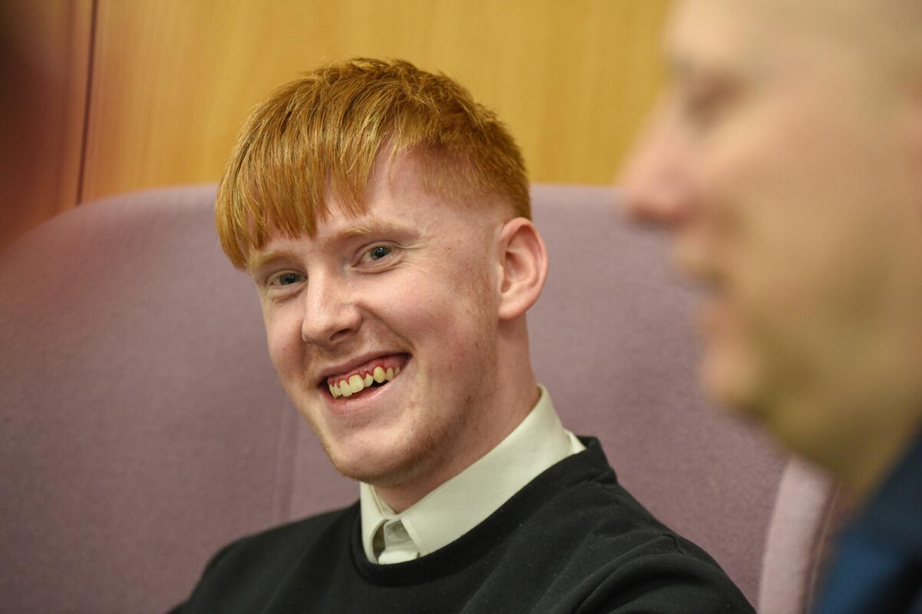 A young man with red hair and a black sweater smiling while sitting on a purple chair indoors.